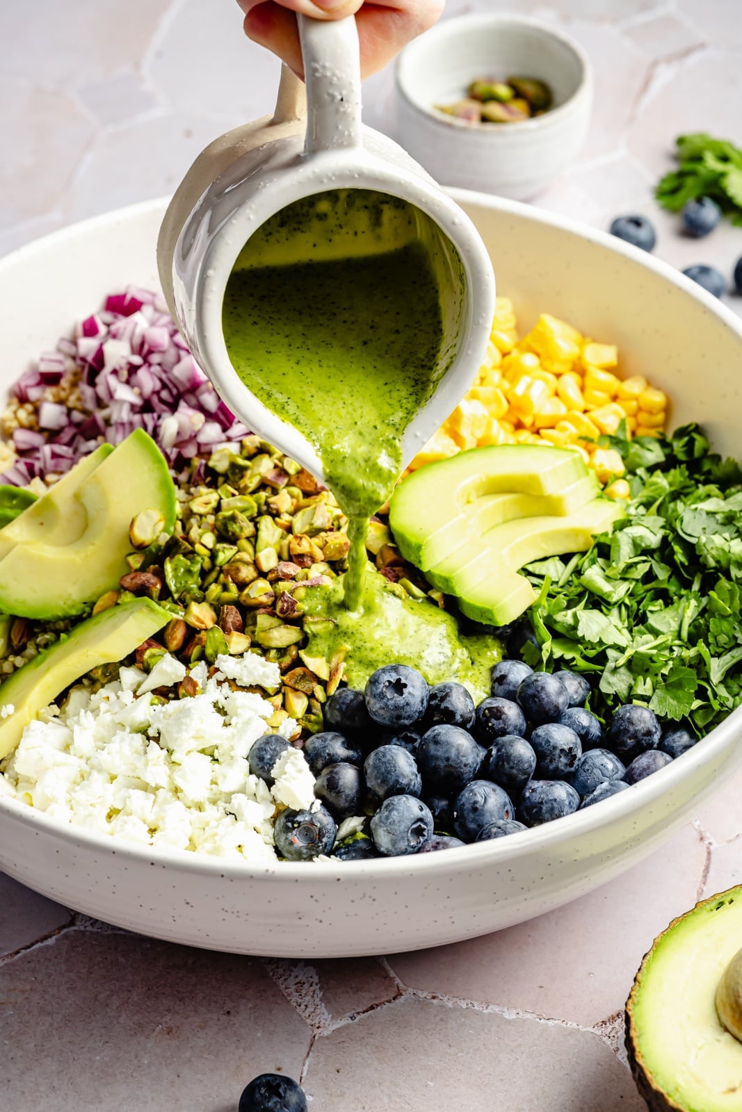 Beautiful avocado blueberry quinoa salad loaded with fresh produce, tangy feta, chopped pistachios, and tossed in a flavorful cilantro lime dressing. This delicious quinoa salad is packed with plant-based protein and is perfect for summer lunches, parties, and picnics. pouring dressing onto a blueberry quinoa salad