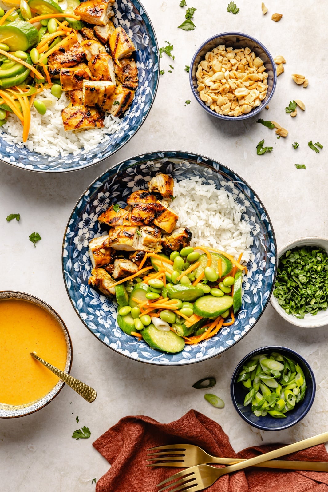 High-protein grilled peanut chicken bowls with edamame cucumber salad and an extra drizzle of flavorful peanut sauce. Assemble these peanut chicken bowls with fluffy rice or quinoa, roasted peanuts, and fresh garnishes for a delicious summer weeknight dinner you'll crave! two grilled peanut chicken bowls next to a bowl of peanut sauce