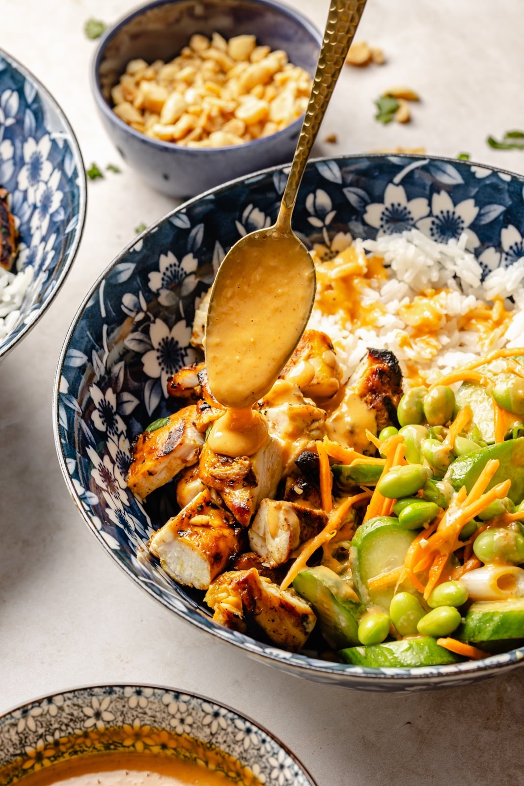 High-protein grilled peanut chicken bowls with edamame cucumber salad and an extra drizzle of flavorful peanut sauce. Assemble these peanut chicken bowls with fluffy rice or quinoa, roasted peanuts, and fresh garnishes for a delicious summer weeknight dinner you'll crave! drizzling peanut sauce onto a peanut chicken bowl