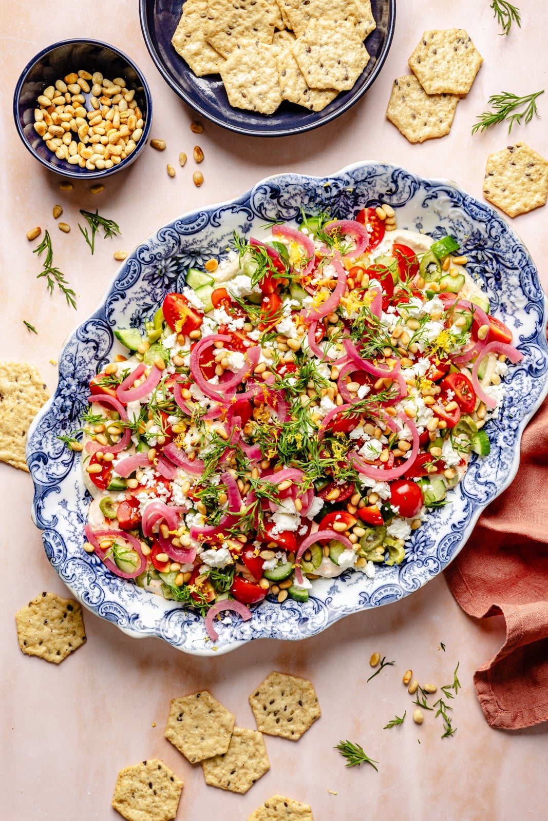 Easy, flavorful Mediterranean dip layered with hummus, creamy yogurt, and fresh toppings like olives, cucumber, and feta cheese. This savory party appetizer is a fun take on classic 7-layer dip and is delicious served with pita chips, pretzels, or naan bread! easy layered mediterranean dip on a platter next to crackers