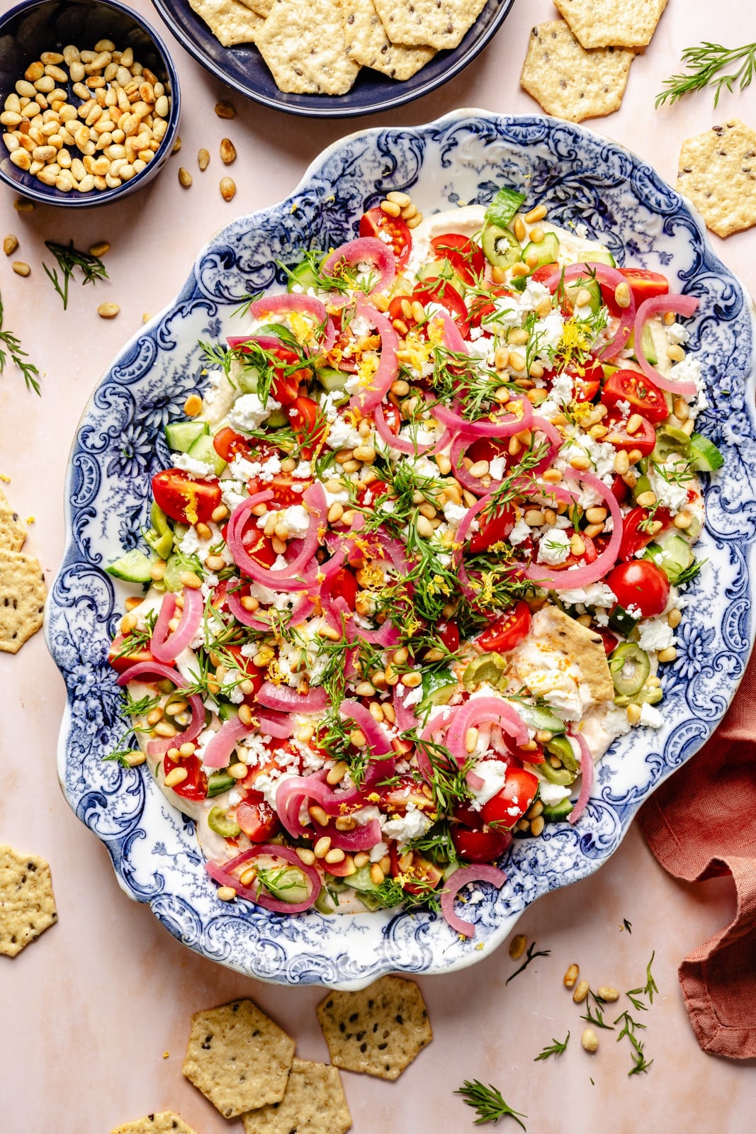 Easy, flavorful Mediterranean dip layered with hummus, creamy yogurt, and fresh toppings like olives, cucumber, and feta cheese. This savory party appetizer is a fun take on classic 7-layer dip and is delicious served with pita chips, pretzels, or naan bread! layered mediterranean dip on a blue platter