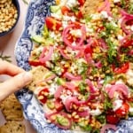 dipping a cracker into a layered mediterranean dip on a platter