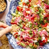 dipping a cracker into a layered mediterranean dip on a platter