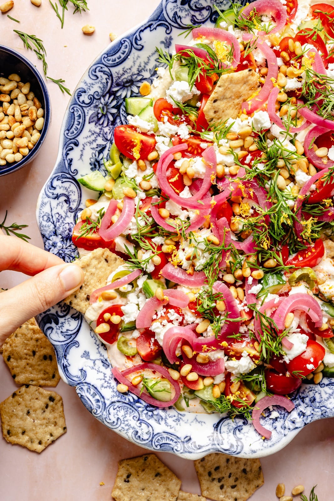 Easy, flavorful Mediterranean dip layered with hummus, creamy yogurt, and fresh toppings like olives, cucumber, and feta cheese. This savory party appetizer is a fun take on classic 7-layer dip and is delicious served with pita chips, pretzels, or naan bread! dipping a cracker into a layered mediterranean dip