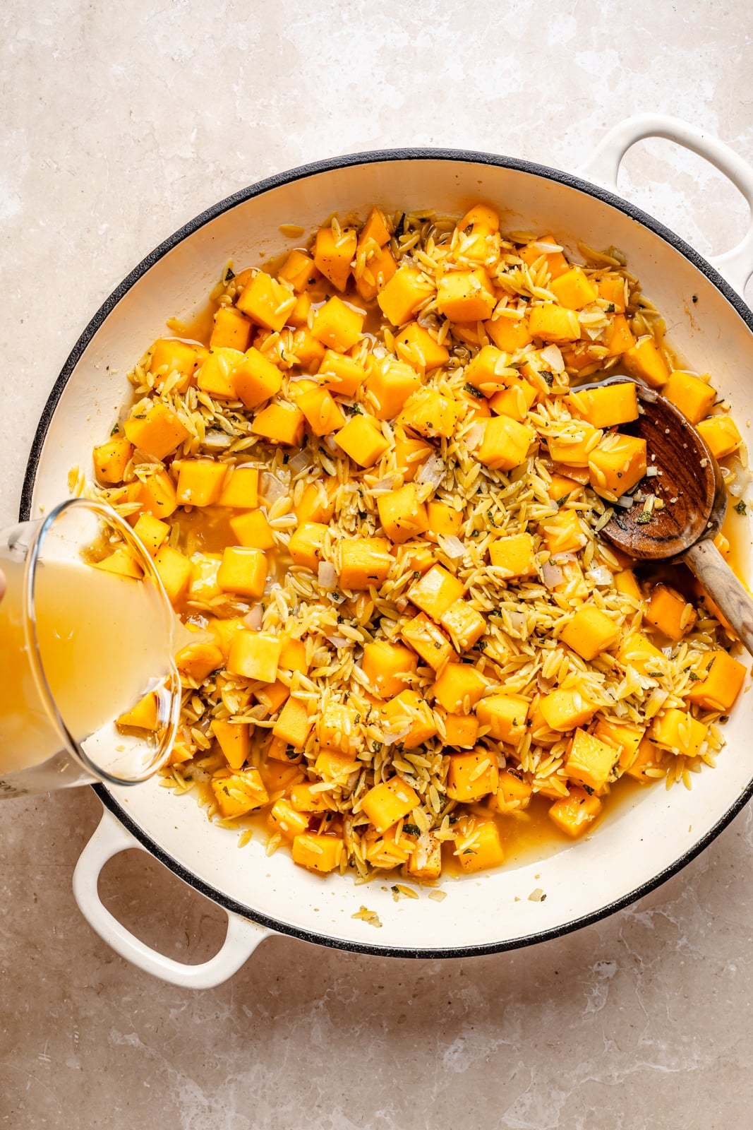 Creamy one pan brown butter sage orzo with spicy sausage, tender butternut squash, and tangy goat cheese. This sweet and savory orzo recipe is filled with cozy fall flavors, plus an extra boost of veggies from spinach! A beautiful weeknight dinner the whole family will love. pouring broth into a pan with orzo and butternut squash