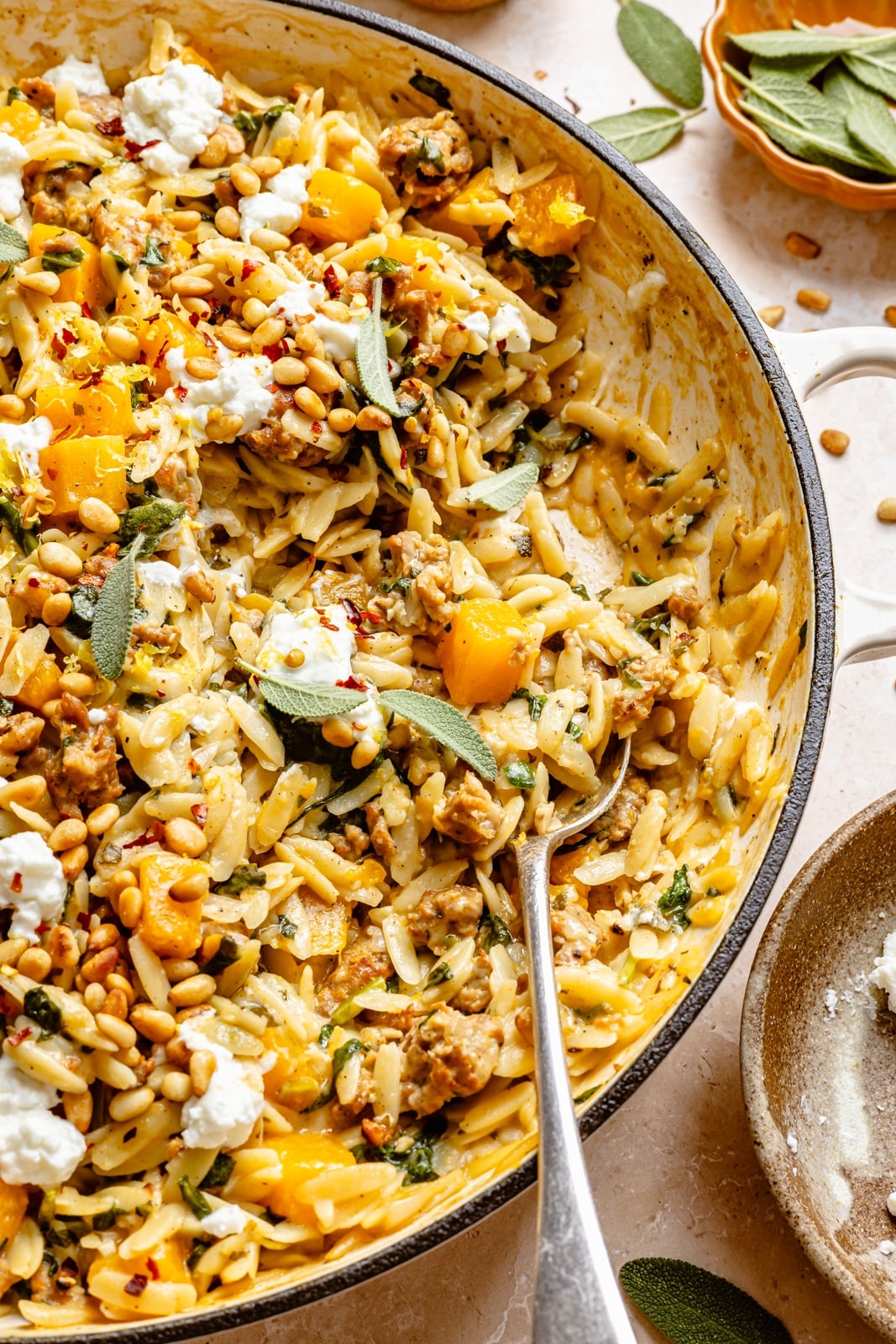 Creamy one pan butternut squash orzo with rich, sage-infused brown butter, spicy sausage, and tangy goat cheese. This sweet and savory orzo recipe is filled with cozy fall flavors, plus an extra boost of veggies from spinach! A beautiful weeknight dinner the whole family will love. one pan butternut squash orzo with sausage in a pan with a spoon